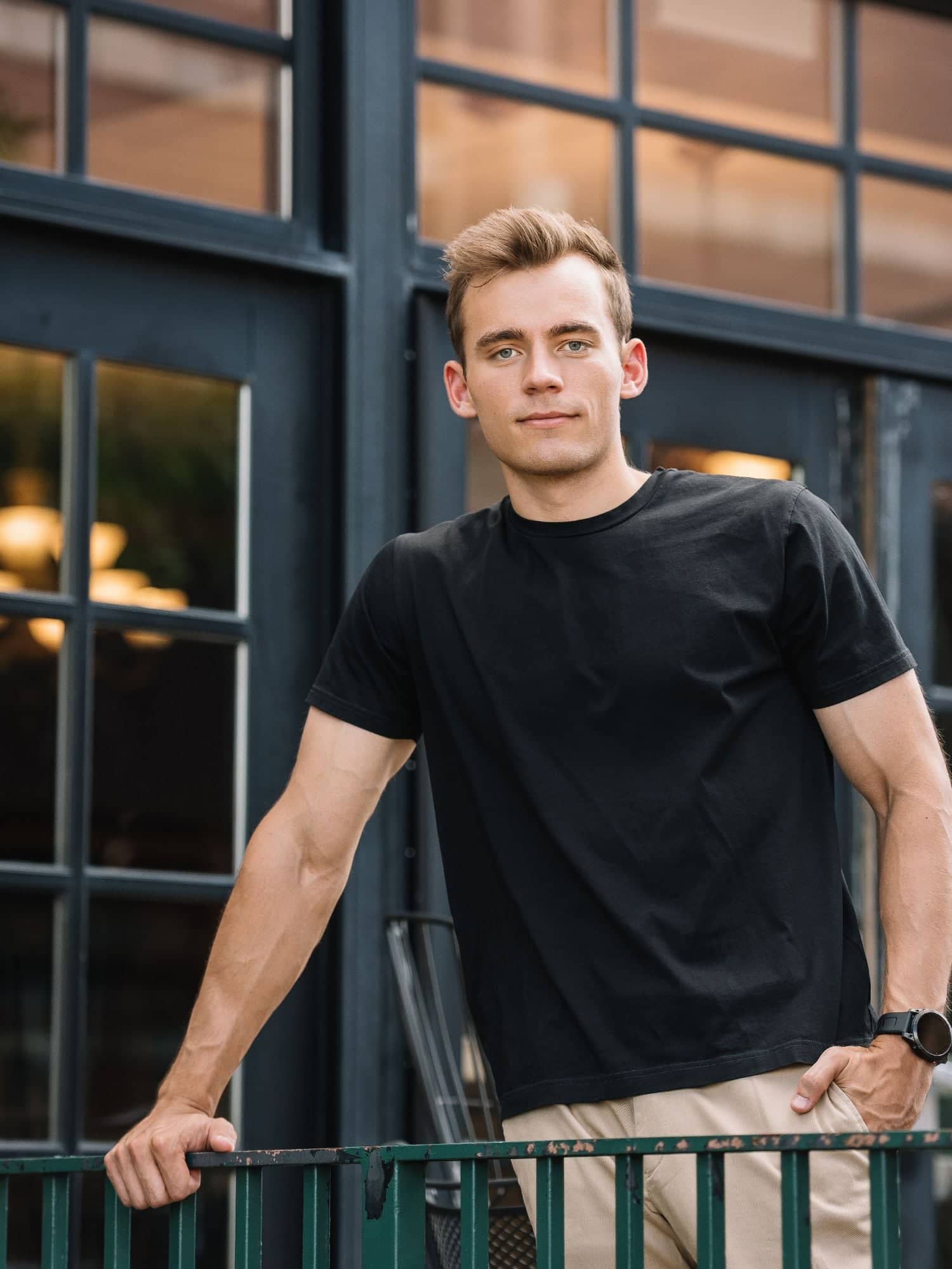 Kyle Wisniewski, in a black T-shirt and khaki trousers, one hand on a green railing, before dark-framed glass doors