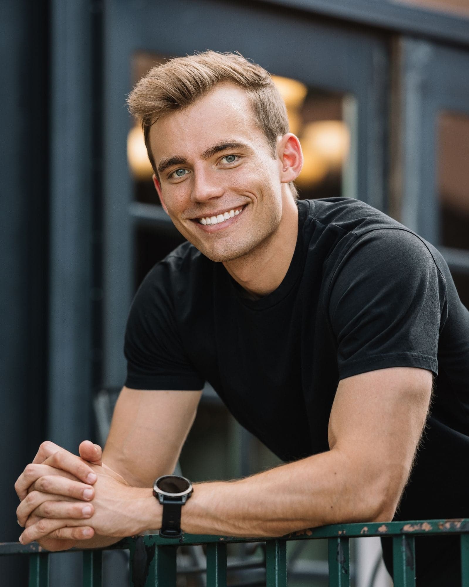Kyle Wisniewski, smiling in a black T-shirt, leaning on a green railing with his hands clasped, dark window frames behind