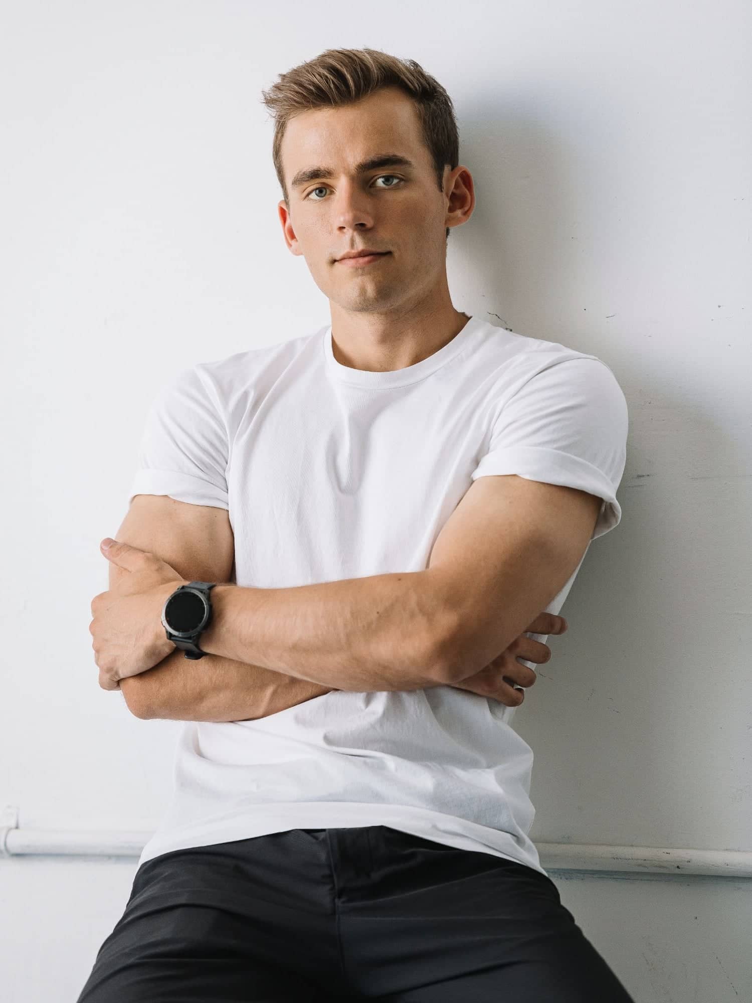 Kyle Wisniewski, in a white T-shirt with his arms crossed and a black watch, leaning against a white wall