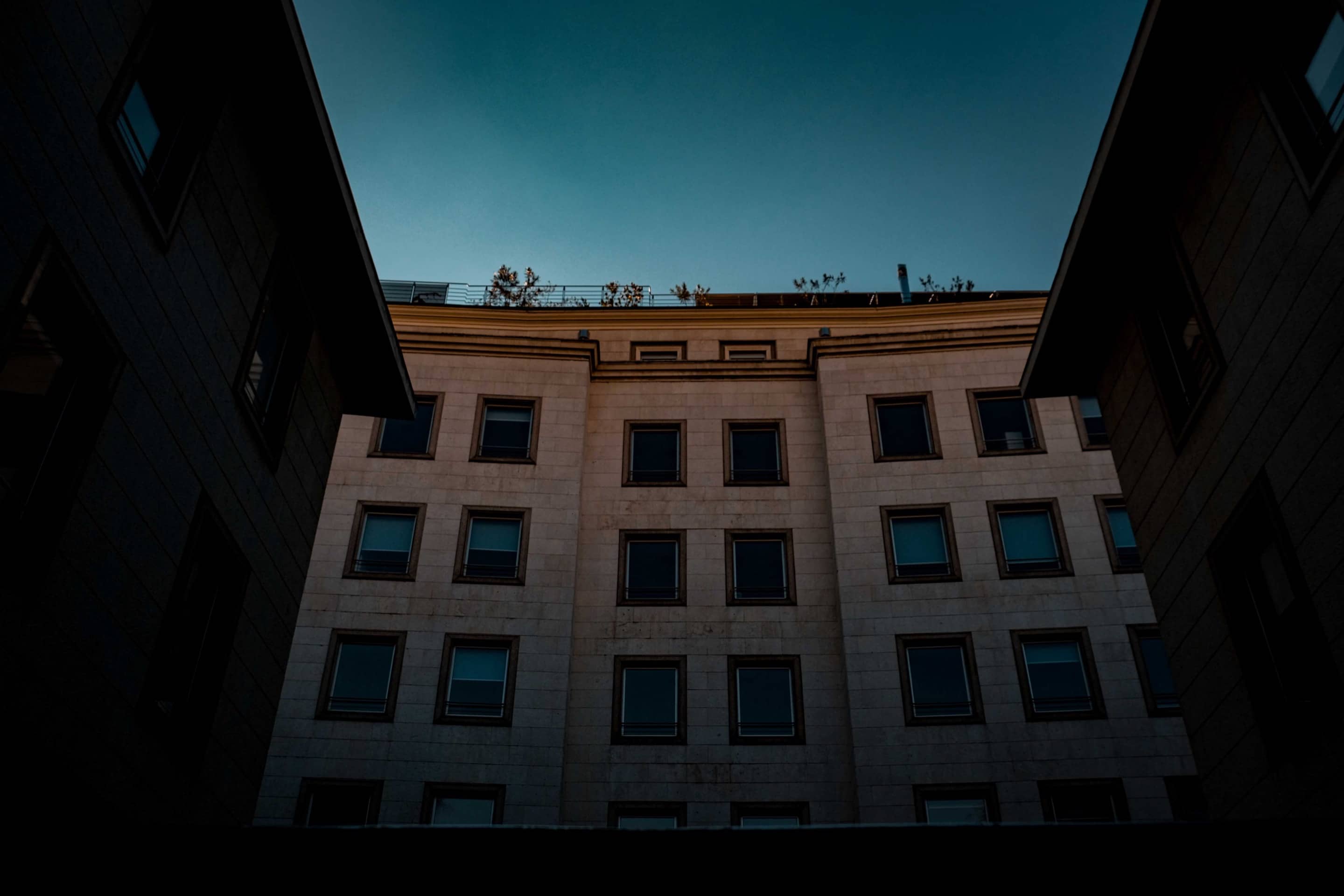 A pale stone building with rows of square windows, seen from below between two dark wings, under a deep blue evening sky.