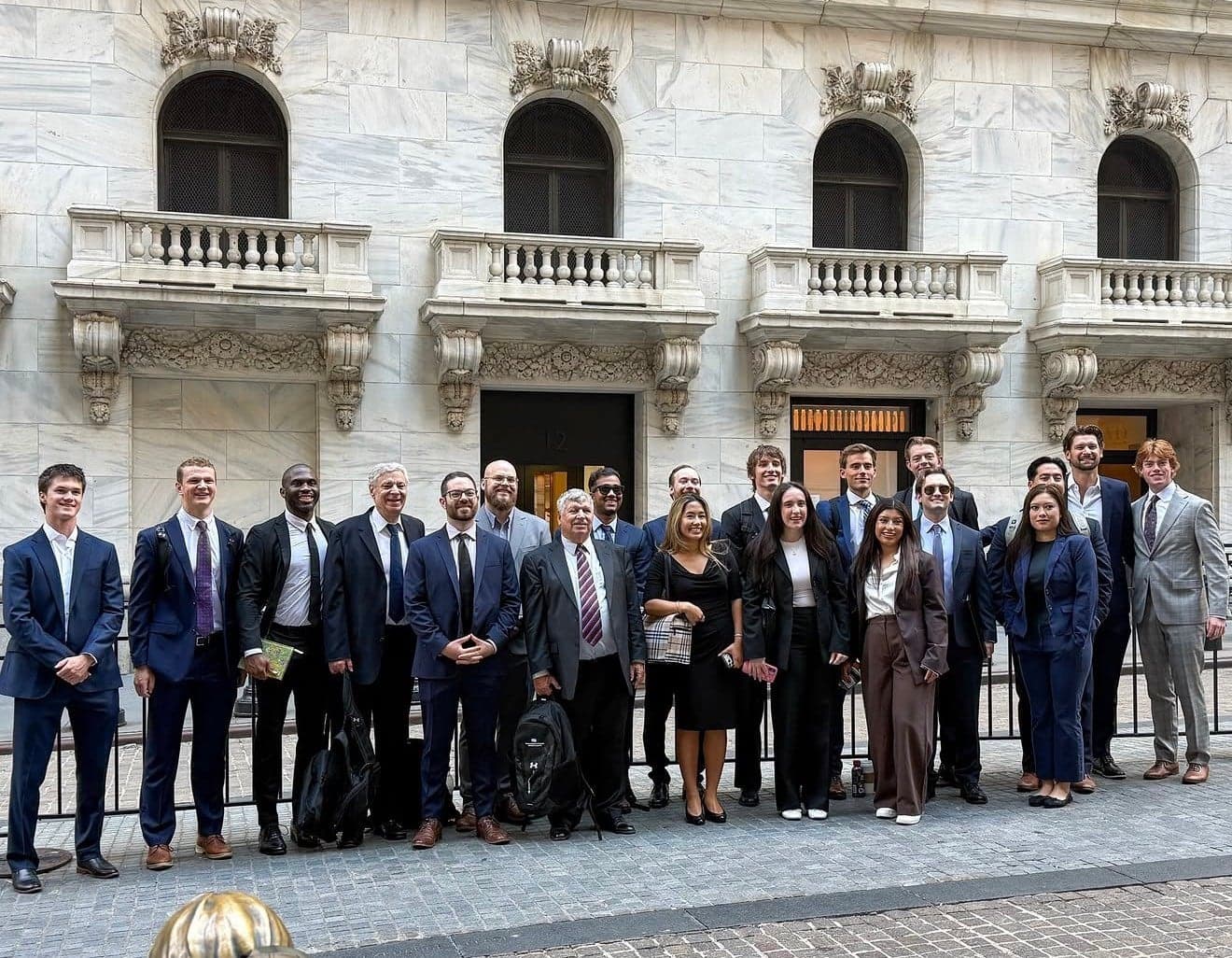 A group in suits and business attire lined up along a black railing outside the marble facade of the New York Stock Exchange, its arched windows and carved stone balconies behind them
