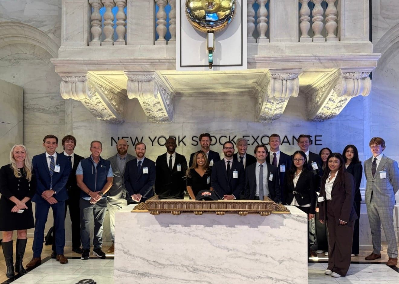 A group in suits standing behind the marble bell podium on the New York Stock Exchange floor, the words NEW YORK STOCK EXCHANGE on the wall and the carved balcony above them, a Denver cap resting on the podium