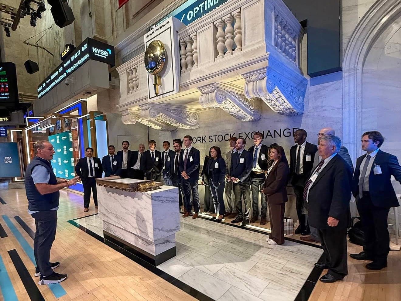 A man in a vest addressing a group in suits beside the bell podium on the New York Stock Exchange trading floor, ticker displays lit overhead