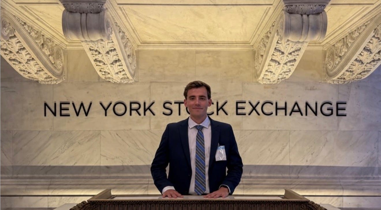 Kyle Wisniewski in a navy suit and striped tie, both hands on the marble bell podium of the New York Stock Exchange, the words NEW YORK STOCK EXCHANGE on the wall behind him